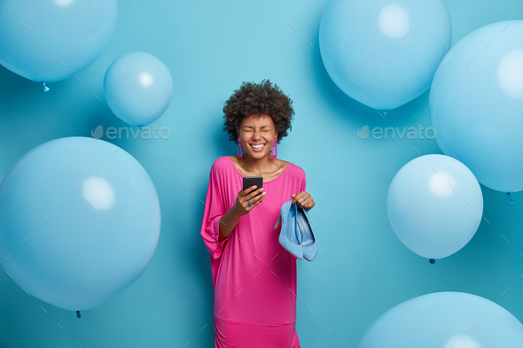 Overjoyed dark skinned woman in pink dress holds mobile phone chats online asks friend for advice wh