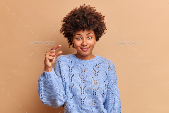 Smiling good looking young woman with curly hair makes size gesture with fingers shapes something sm