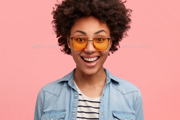 Close up shot of female hipster with black skin, curly hair, smiles  positively, wears shades and den Stock Photo by wayhomestudioo