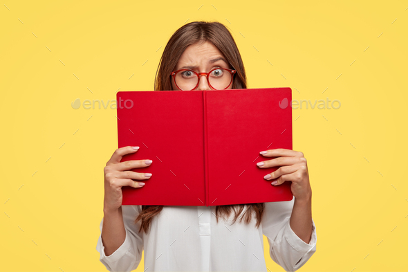 Puzzled dark haired woman raises eyebrows, looks with puzzlement over red textbook, wears white shir