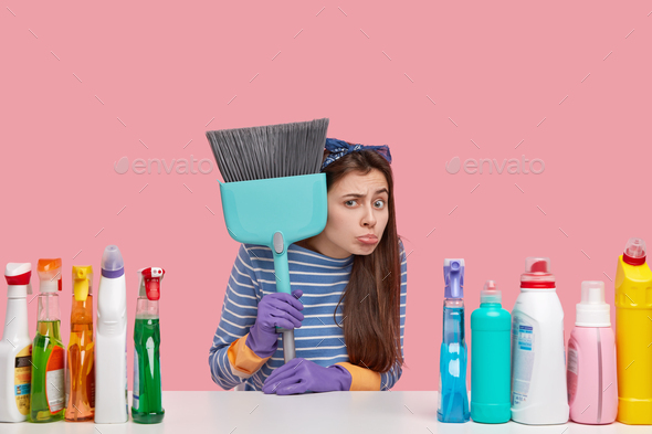 Unhappy young cleaning service employee holds broom, uses numerous cleaning substances, arent ready
