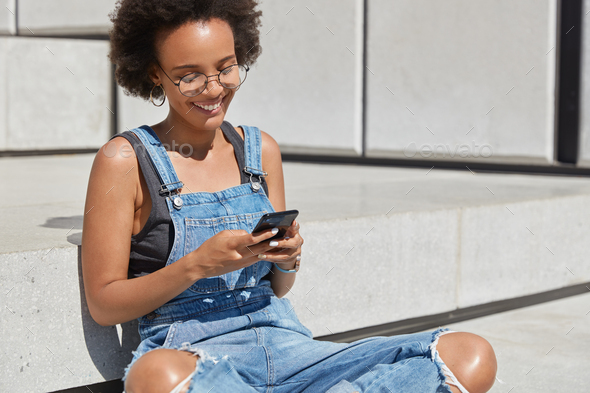 Happy fashionable feminine blogger sits crossed legs outside, texts message for post on personal web