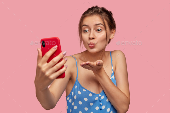 Pleasant looking girlfriend flirts with man over smart phone, makes air kiss during video call, hold