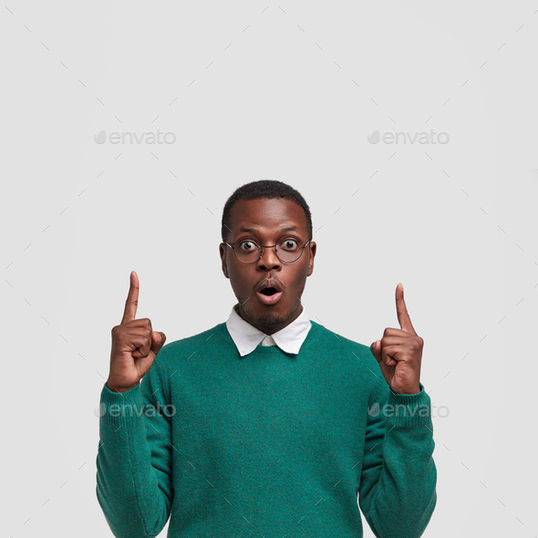 Young black young man has stupefied expression points upwards with both index fingers, has bated bre