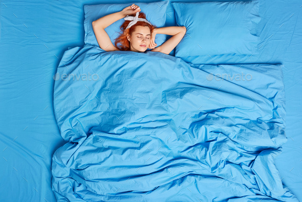 Top view of calm redhead woman sleeps peacefully in bed enjoys sweet dreams keeps eyes closed rests