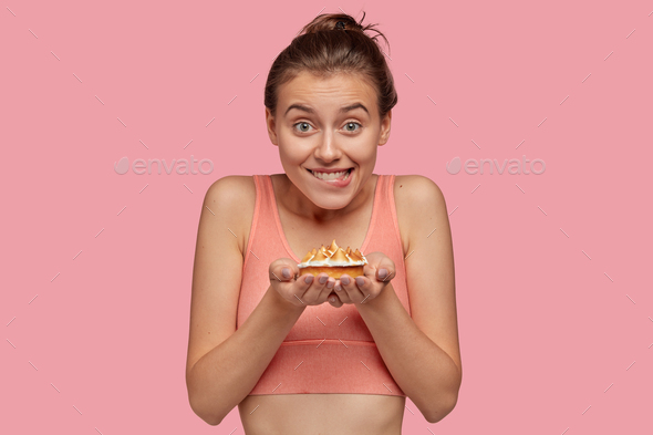 Photo of cheerful fitness lady feels temptation while holds delicious sweet cake, wears casual top,
