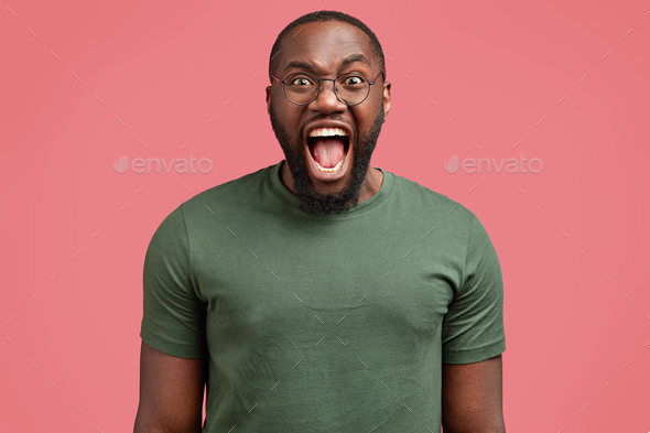 Studio shot of mad African American male screams angrily at someone, being irritated, wears casual t