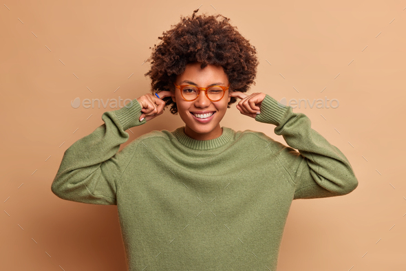 Positive dark skinned curly haired woman smiles happily plugs ears avoids very loud music wears tran
