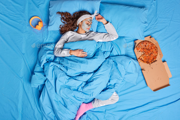 Top view of relaxed woman fall asleep with clay mask on face sees sweet dreams at home enjoys eating
