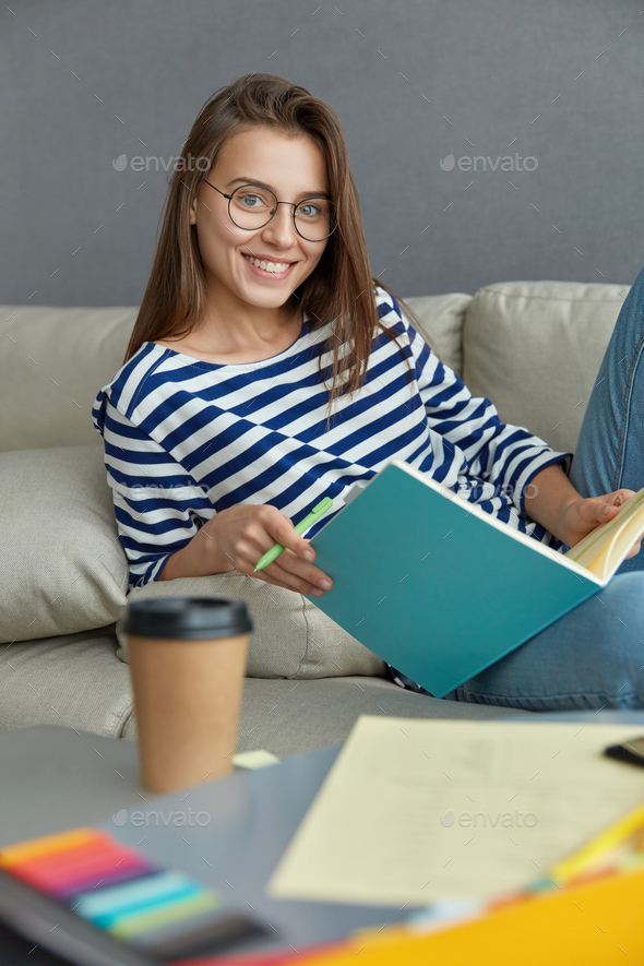 Vertical shot of cheerful brunette woman checks course work, reads text information, from textbook,