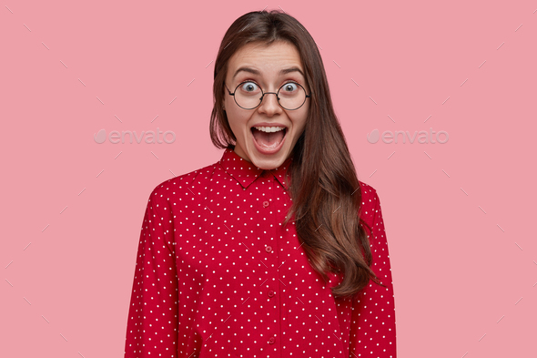 Attractive brunette overjoyed woman opens mouth and yells from joy, wears pink polka dot blouse, spe