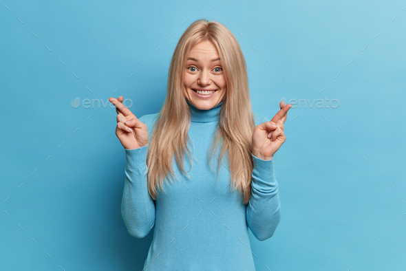 Cheerful smiling blonde woman waits for special moment keeps fingers crossed wishes good luck dresse