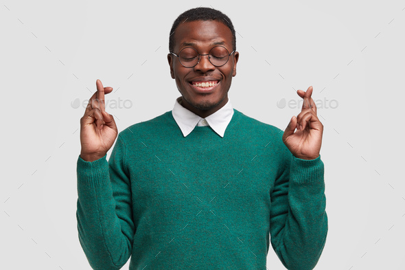 Smiling Afro American man prays for fullfilment of his wish, crosses fingers, wishes good luck, wear