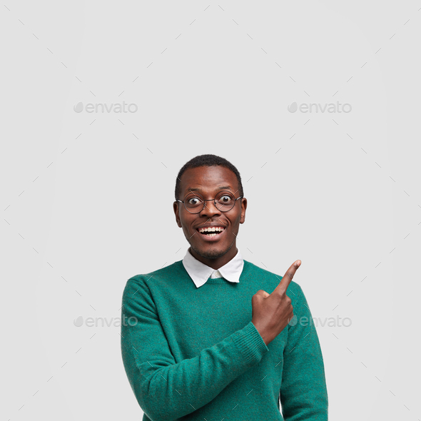 Cheerful smiling dark skinned male points with index finger upwards, glad to demonstrate free space
