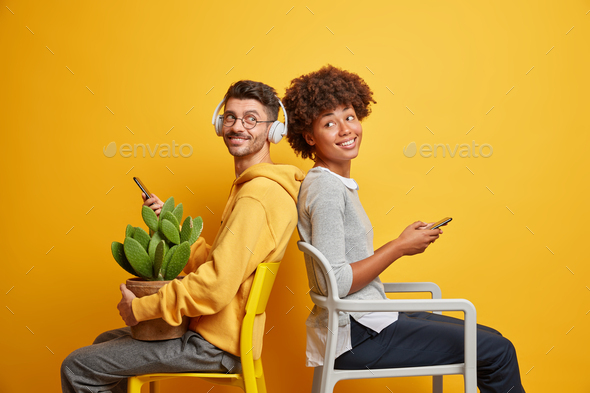 Positive teenage female and male addicted to modern technologies sit back to each other on chairs ho - Stock Photo - Images
