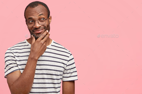 Horizontal shot of attractive dark skinned male with satisfied expression holds hand above chin, loo