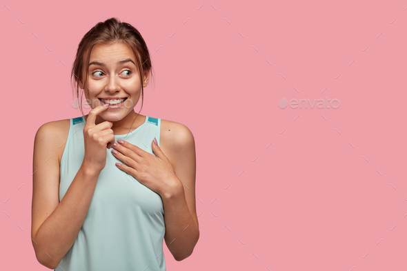 Isolated shot of positive European woman bites finger, keeps hand on chest, smiles positively, looks