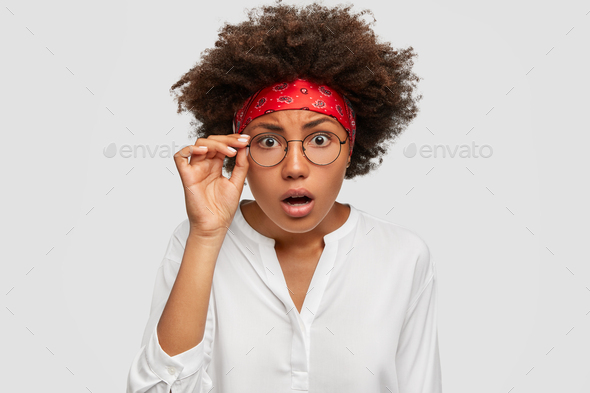 Photo of terrified beautiful woman keeps hand on rim of spectacles, stares with scrupulous look at c