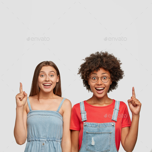 Indoor shot of attractive young females have pleased facial expressions, point upwards, stand closel