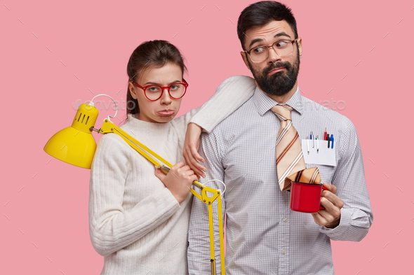 Sad displeased woman and man in spectacles, do common work, hold lamp and mug of coffee, express neg