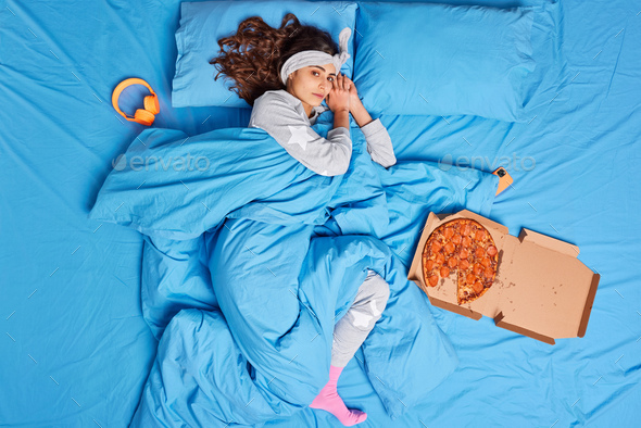 Lovely brunette woman rests on comfortable bed wakes up late dressed in soft nightwear eats pizza be