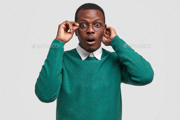 Indoor shot of stupefied black man with amazed facial expression, keeps hand on rim of spectacles, d