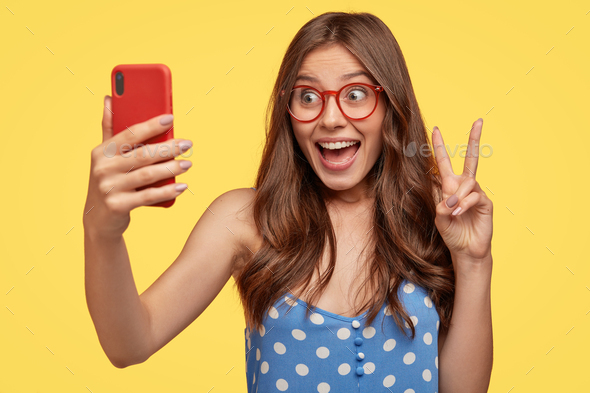 Attractive glad European woman with long hair, makes peace sign, holds mobile phone, poses for selfi