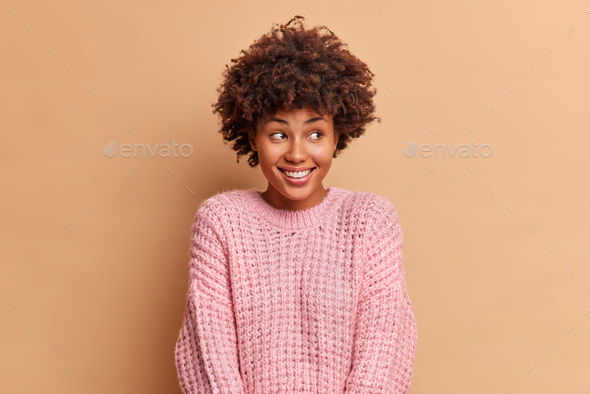 Photo of glad millennial dark skinned woman with Afro hair smiles gently and looks away being in hig