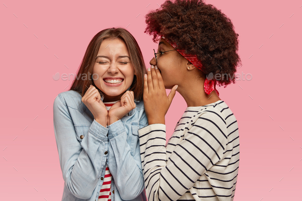 Black ethnic woman whispers secret to her Caucasian female with toothy smile, gossip together, discu