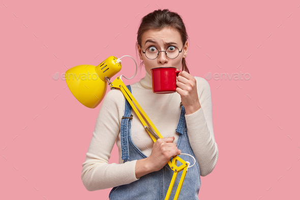Photo of puzzled Caucasian woman raises eyebrows, drinks beverage from red mug, has break after stud