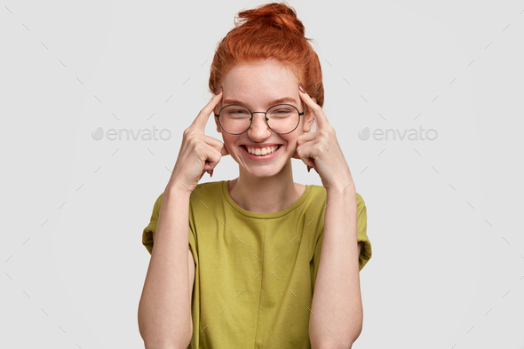 Use your brain. Happy red haired girl holds both index fingers on temples, tries to think before act
