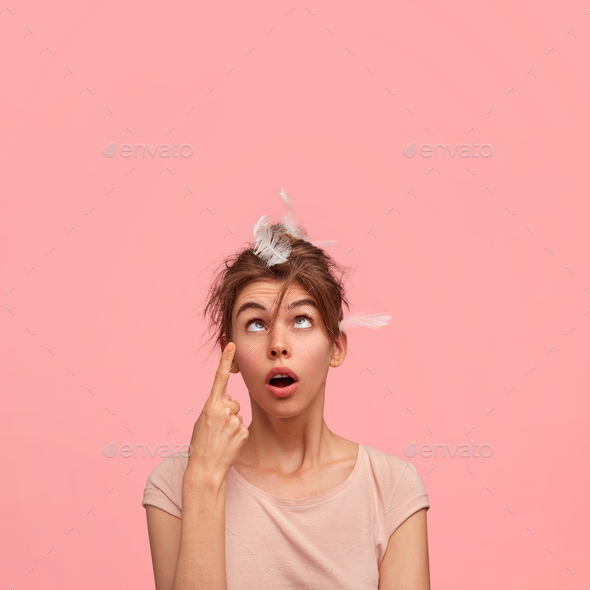 Crazy sleepy young woman crosses eyes, has feathers on head, points upwards, has comic expression, w