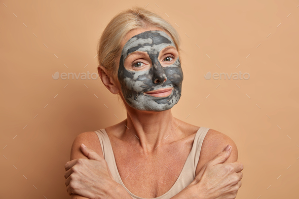 Headshot of lovely middle aged woman applies clay mask on face looks with satisfied expression at ca