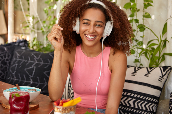 Happy Afro American female meloman listens music with high volume in headphones, sits in cafeteria,