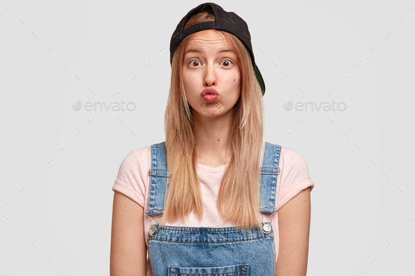Gorgeous funny female college student keeps lips round, pouts lips, wears trendy cap and overalls, s