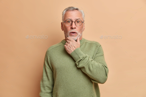 Surprised bearded grey haired speechless man holds chin and stares bugged eyes hears secret or shock