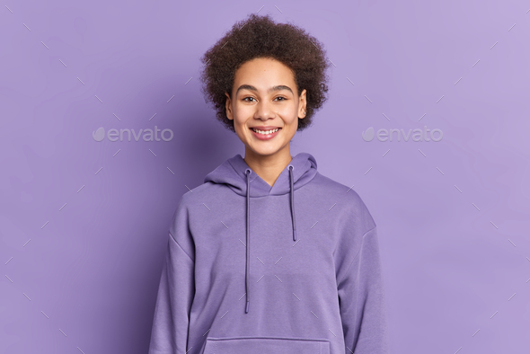 Happy ethnic teenager with Afro hair smiles positively wears purple hoodie being in good mood poses
