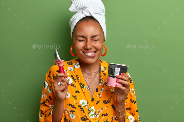 Studio shot of happy Afro American lady enjoys eating tasty ice cream with spoon bites lips and clos