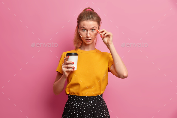 Impressed young European woman looks very surprised through big spectacles holds breath drinks takea