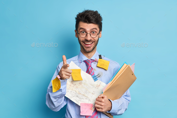 Cheerful young man coworker happy to finish project work covered with papers and stickers points at
