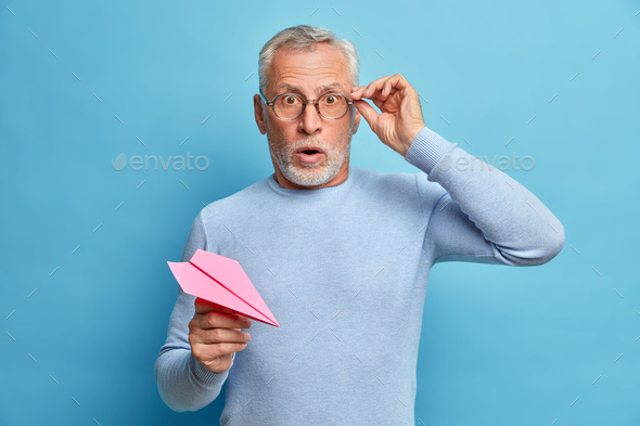 Isolated shot of grey haired man holds breath keeps hands on spectacles being frightened holds paper