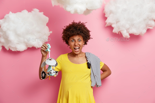Angry future mother holds mobile toy shouts irritated dressed in yellow dress has big abdomen carrie - Stock Photo - Images