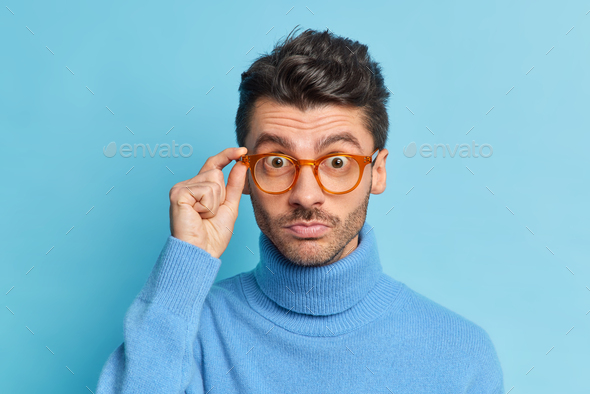 Close up shot of surprised Caucasian guy with thick bristle keeps hand on rim of spectacles wears ca