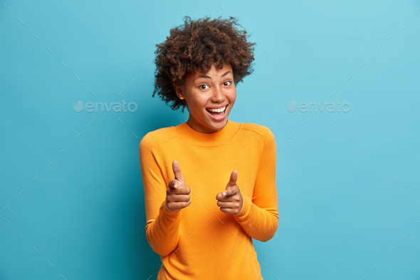 You got it. Happy young curly Afro American woman with curly hair points at camera with index finger