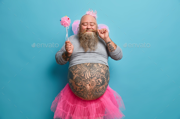 Positive funny bearded male princess holds magic wand and entertains guests on costume party shows f