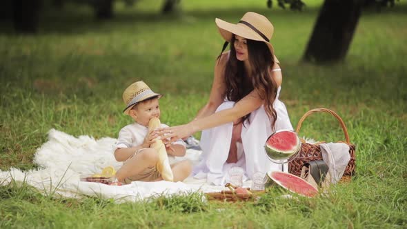 Young Beautiful Woman Give a Baguette To Her Child at a Picnic in the Park in the Summer