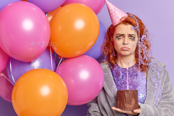 Indoor shot of unhappy ginger European woman purses lips going to cry celebrates birthday alone hold
