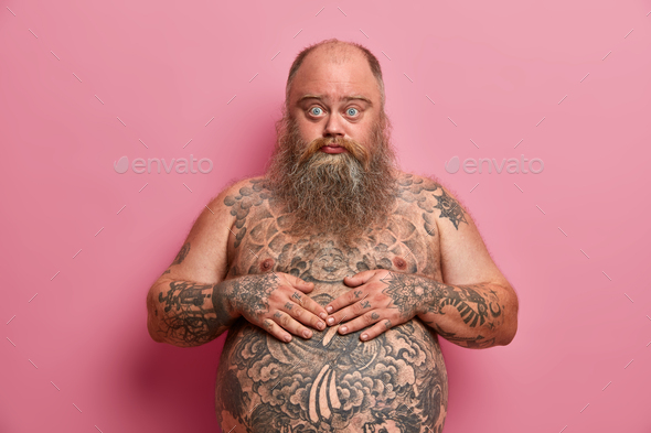 Surprised handsome blue eyed fatso man keeps hands on tattooed belly looks with embarrassed expressi