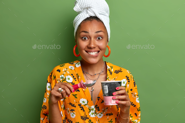 Pretty smiling woman has dark skin bites lips with appetite as eats tasty strawberry ice cream spend