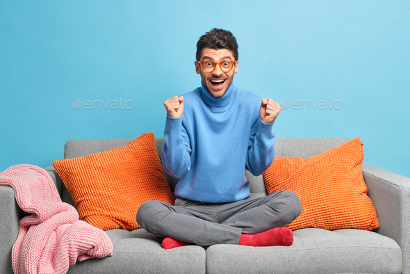 Emotional European man sits crossed legs on sofa with cushions watches  sport game supports favorite Stock Photo by wayhomestudioo